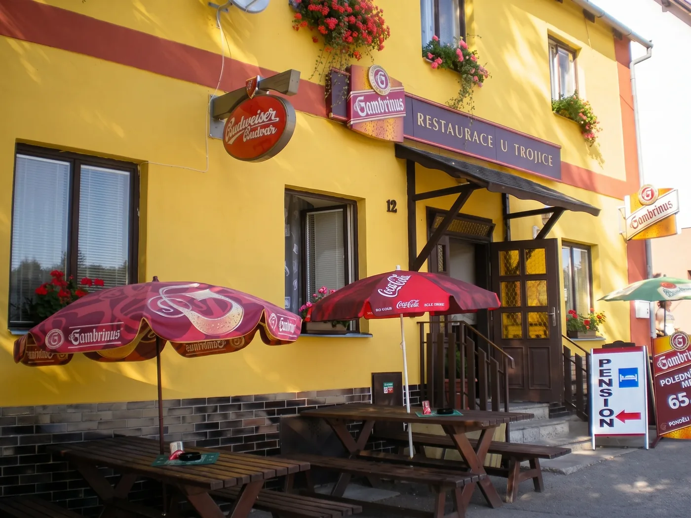 Vchod do restaurace U Trojice s cedulí nad markýzou, slunečníky a stoly na terase před domem